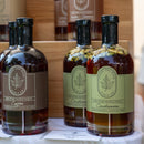 Three bottles of Under The Bark Syrup Co Cardamom Maple Syrup sit on a table before a wooden shelf, with visible cardamom pods floating inside the syrup.