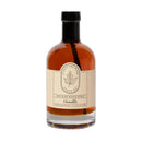 A clear glass bottle of Under The Bark Syrup Co Vanilla Maple Syrup with a black cap, featuring amber syrup and a label with a maple leaf and product information, showcasing its rich vanilla blend.