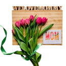 A bouquet of pink tulips with a green ribbon rests on the Words with Boards, LLC Large Personalized Cutting Board (12.5" x 16") engraved with “WE LOVE MOMMY,” placed beside a colorful card reading “MOM.”.