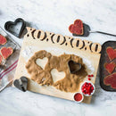 Heart-shaped cookies are made on the Words With Boards Large Personalized Cutting Board (12.5" x 16") by Words with Boards, LLC. Cookie cutters, red sprinkles, a wooden "XOXO" sign, and finished cookies on a tray sit atop a marble countertop.