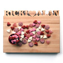 The Words with Boards Personalized Cutting Board Small 9.5" x 12.5" by Words with Boards, LLC features “RICKI ♥ ALAN” cut out at the top and holds heart-shaped candies—making it a charming kitchen gift for any sweet occasion.