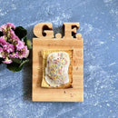 A Words With Boards Gluten Free Personalized Cutting Board (6 1/4" x 9 1/2", by Words with Boards, LLC), engraved with "G.F.," holds a sprinkle-topped pastry. A small pink-flowered plant sits to the left on a textured blue surface.