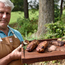A man in a brown apron holds a Words With Boards Personalized Meat Cutting Board with Smart Slope Design by Words with Boards, LLC, engraved with "CARNIVORE," displaying grilled meats, garlic, and herbs outdoors among lush greenery.