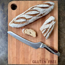 A serrated bread knife and rustic bread rest on the Words With Boards, LLC Seriously GLUTEN FREE Cutting Board Large 16” x 12.5”, featuring a flour-dusted crust, ideal for gluten free food prep and separation.