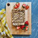 The Words With Boards, LLC Seriously Gluten Free Cutting Board Large 16” x 12.5” holds chocolate-drizzled cookie bars and strawberries. A yellow checkered cloth to the left on a blue surface ensures ideal food separation.