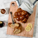 A loaf of braided challah bread, partially sliced, rests on the Words With Boards Seriously Kosher Cutting Board for Milk (16” x 12.5”) by Words with Boards, LLC, surrounded by a striped towel, honey dipper, knife, lemon slices, and a glass with lemon.