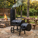 A Yoder Smokers Durango 20 Offset Smoker by Yoder Smokers stands on a stone patio near a fire pit, smoke drifting from its chimney. A picnic table behind is set with food, ideal for BBQ gatherings surrounded by lush trees.