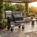 A large black and silver Yoder Smokers YS640S Competition Pellet Grill with Wire Shelves is on wheels and sits on a stone patio in an outdoor kitchen, surrounded by greenery, string lights, benches, and a fire pit at sunset.