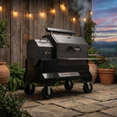 A Yoder Smokers YS640S Competition Pellet Grill with Wire Shelves stands on a stone patio near a wooden wall with string lights, surrounded by potted plants and greenery at sunset. Smoke rises from the grill, indicating it is in use.
