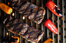 Two grilled steaks with char marks cook alongside red, orange, and yellow mini bell peppers over direct flame on a Yoder Smokers YS640 grill, using the Yoder Smokers YS640 Two-Piece Heat Diffuser glowing warmly below.