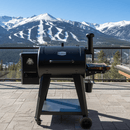 A BBQ Boards® Pit Boss 700DX Side Cutting Board is attached to a black pellet grill on a stone patio, overlooking forested mountains and snow-covered ski slopes beneath a clear blue sky.
