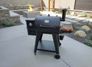 A black Pit Boss 700DX outdoor pellet grill features a BBQ Boards® Pit Boss 700DX Side Cutting Board as a side shelf and a bucket underneath, set on a patio with a modern fire pit and stylish landscaping in the background.