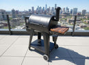 A BBQ Boards® Pit Boss 700DX Side Cutting Board is attached to a black pellet grill on a rooftop patio, with the city skyline and skyscrapers visible under a sunny sky.