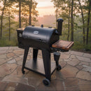 A BBQ Boards® Pit Boss 700DX Side Cutting Board is attached to a black pellet grill with smoker box, standing on a stone patio with sunlight streaming through trees in the background.