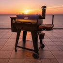 A BBQ Boards® Pit Boss 700DX Side Cutting Board is mounted on a black outdoor smoker grill on a tiled patio, overlooking calm water at sunset with an orange sky in the background.