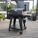 A black Pit Boss 700DX pellet grill with a side shelf and chimney stands on a modern patio, featuring a BBQ Boards® Pit Boss 700DX Side Cutting Board from BBQ Boards® attached to the side board, surrounded by outdoor seating and potted plants.