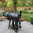 A BBQ Boards® Pit Boss 700DX Side Cutting Board is attached to a black pellet grill on a patio, surrounded by patio furniture, potted plants, and blooming bushes in a landscaped backyard with a wooden fence.