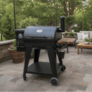 A BBQ Boards® Pit Boss 700DX Side Cutting Board is installed on a black Pit Boss 700DX pellet grill smoker, set on a patio with outdoor furniture, greenery, and string lights hanging above.