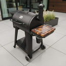 A Pit Boss 700DX smoker grill with a black BBQ Boards® Side Cutting Board by BBQ Boards® attached stands on a tiled patio near tall windows and a planter with grass-like green plants.