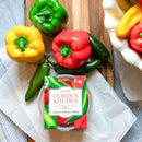 A 2-pack of In The Curious Kitchen Jalapeño Pimento Cheese, a Southern-style cheese spread, sits on parchment paper beside vibrant bell peppers and green jalapeños on a wooden surface.