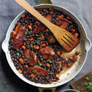 A skillet with sautéed black beans, sliced sausage, diced carrots, and chopped parsley, seasoned with SALTOPIA Infused Sea Salts' BBQ Blend: HE MAN, with a wooden spatula on top—all set on a gray fabric surface.