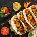 Three shrimp tacos with avocado, corn, tomato, and herbs are arranged on a board and seasoned with SALTOPIA Infused Sea Salts’ SHE DEVIL Spicy Herbal Blend. Nearby are a lemon half, lime wedge, whole shrimp, tomato slice, and salsa bowl.