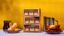 Three Spicewalla 3 Pack Signature Sugar Collection boxes from Spicewalla are stacked between honeycomb, syrup, and French toast on purple plates, with a flour sifter in the background and a yellow tiled wall.
