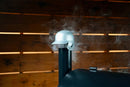 A silver Pimp My Grill® Baseball Batting Helmet BBQ Chimney Cap sits on a grill with smoke rising from it.