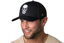 A smiling man wears the Haka Hat Skull Trucker Hat in black, featuring a white skull graphic on the front, and a beige t-shirt, set against a white background.