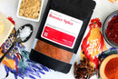 A package labeled World Spice Rooster Spice from World Spice, ideal for infusing a bold kick of chili powder into your favorite Southeast Asian dishes, sits surrounded by star anise, salt, ground ginger, and crushed red pepper—all artfully displayed on a tablecloth adorned with vibrant rooster illustrations.