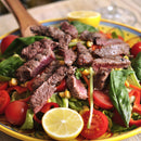 A vibrant salad topped with sliced grilled steak seasoned with SALTOPIA Infused Sea Salts’ HOLY MOLY Taco Seasoning, cherry tomatoes, spinach, corn, and lemon halves, served on a colorful plate.