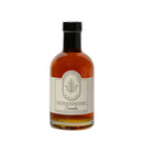 A clear glass bottle of amber Smoke Maple Syrup by Under The Bark Syrup Co, featuring a black cap and white label with maple leaf illustration—perfect for cocktails.