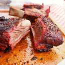 Close-up of juicy, smoked beef ribs with a charred crust, sliced on a wooden board and finished with SALTOPIA Infused Sea Salts BBQ Blend: HE MAN, as savory meat juices glisten on the surface.