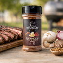 A jar of Texan Heartland Smokehouse Beef & Steak Seasoning, celebrated for its robust smokehouse flavor, is placed on a wooden surface next to sliced grilled steak, a whole garlic bulb, red onion, assorted spices, and with an outdoor grill softly blurred in the background.