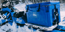 A blue cooler secured to the back of a blue snowmobile with a Blue Coolers Secure Universal Tie Down Kit For Adventurers in a snowy forest.