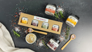 A variety pack of four small and three full-size jars of B.T. Leigh’s Garlic & Rosemary Finishing Salt, displayed with scattered salt crystals, rosemary sprigs, and wooden tasting spoons on a dark surface.