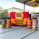 The Chiles & Smoke Sonoran BBQ Starter Box by Chiles and Smoke—featuring four seasonings and a BBQ sauce inspired by the authentic flavors of the Sonoran Desert—is displayed on an outdoor table, ideal for live-fire cooking, with houses and a patio cover softly blurred in the background.