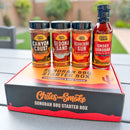 Four spice jars and a BBQ sauce bottle rest atop the Chiles & Smoke Sonoran BBQ Starter Box by Chiles and Smoke, displayed outdoors on a table with plants and a fence softly blurred in the background. This set is ideal for anyone looking to experience genuine Sonoran Desert flavors in their live-fire cooking.
