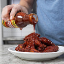 A person pours Chiles & Smoke Smoky Sonoran BBQ Sauce from a Chiles and Smoke bottle onto a plate of cooked chicken wings on a kitchen counter.