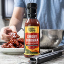 A bottle of Chiles and Smoke Smoky Sonoran BBQ Sauce rests on a counter. In the background, someone coats chicken wings with the smoky sauce from a bowl. Metal tongs and a plate of wings can also be seen.