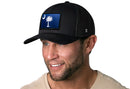 A man with light facial hair wears a beige shirt and the Haka Hat South Carolina Flag Trucker Hat, a black mesh snapback featuring a crescent moon and palmetto tree patch symbolizing the state flag.