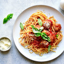 Plate of spaghetti with meatballs in tomato sauce, seasoned with SALTOPIA Infused Sea Salts’ Italian Herbs & Garlic Salt: KITCHEN'S KEEPER, topped with fresh basil and grated cheese, next to a small bowl of grated cheese on a light gray surface.