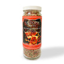 A clear jar of SALTOPIA Infused Sea Salts "Spicy Herbal Blend: SHE DEVIL" displays a colorful mix of pink Himalayan salt, sun-dried tomato seasoning, and herbs. The label features bruschetta topped with tomatoes and herbs.