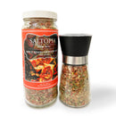 A jar labeled SALTOPIA Infused Sea Salts "Spicy Herbal Blend: SHE DEVIL" stands next to a clear grinder filled with a zesty herbal and sun-dried tomato seasoning blend.