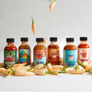 Six bottles from the Culture Hot Sauce Gift Sampler Pack, each with colorful labels, are arranged in a row among light yellow chili peppers, some of which appear to float above them on a plain white background. This vibrant sampler set from Culture Hot Sauce makes the perfect gift and features several of their bestselling hot sauces.