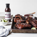 A tray of glazed ribs with a brush sits beside a bowl of sweet heat sauce, a bottle of Terrapin Ridge Farms Raspberry Peach Chipotle Sauce, and lime wedges on a white table with a striped napkin in the corner.