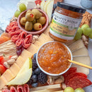 A charcuterie board with cheeses, salami, fresh fruits, pistachios, green olives, and a bowl of Terrapin Ridge Farms Pear Pistachio Jam is served as a gourmet appetizer. A jar of Terrapin Ridge Farms Pear Pistachio Jam is also displayed.