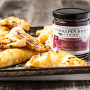 A jar of Terrapin Ridge Farms Hot Pepper Berry Bacon Jam sits beside golden, flaky ham and cheese pastries on a patterned tray atop a rustic wooden table.