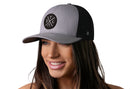 A woman with long brown hair smiles wearing the Haka Hat St. Louis Trucker Hat – a gray and black STL X Snapback by Haka Hat, featuring a round black patch with white lettering, against a plain white background.