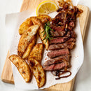 Medium-rare steak with caramelized onions, served with potato wedges dusted in SALTOPIA Infused Sea Salts’ Saltopia Italian Herbs & Garlic Salt: KITCHEN'S KEEPER, plus rosemary and lemon, on parchment atop a wooden board.